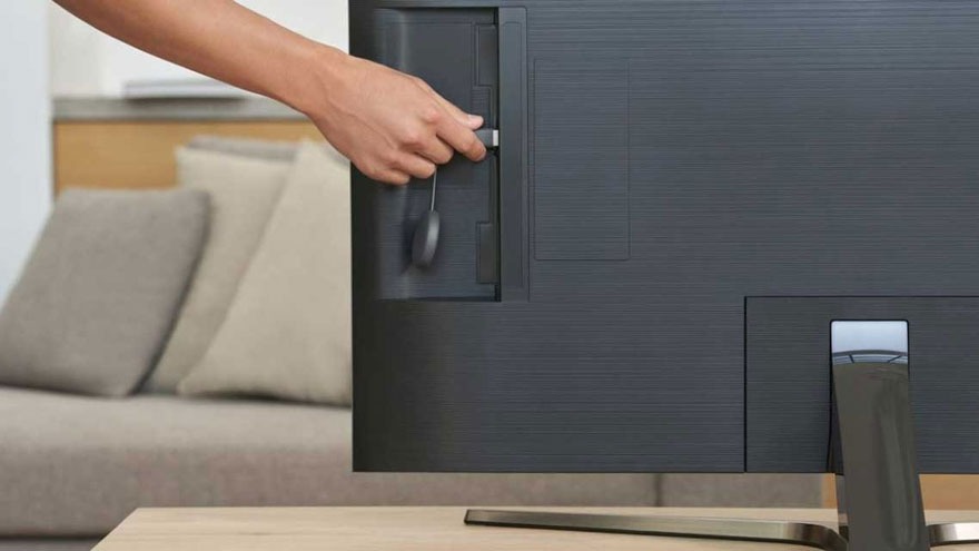Een hand steekt een ronde, zwarte Chromecast in een zwarte televisie. Op de achtergrond staat een beige sofa.