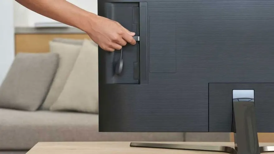 Een hand steekt een ronde, zwarte Chromecast in een zwarte televisie. Op de achtergrond staat een beige sofa.
