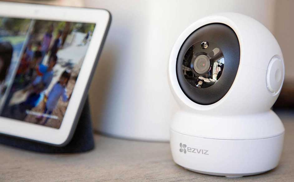 Een witte Ezviz beveiligingscamera staat naast een tablet op een houten tafel. De tablet toont een onscherp beeld.
