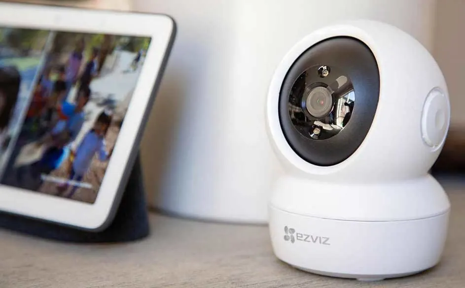 Een witte Ezviz beveiligingscamera staat naast een tablet op een houten tafel. De tablet toont een onscherp beeld.