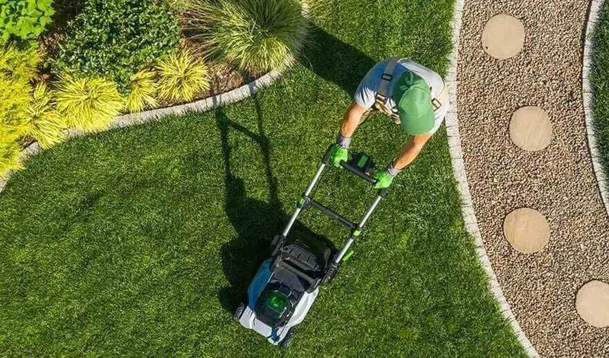 Une personne avec une casquette et des gants verts tond la pelouse près d’un chemin avec des pierres rondes. Marque : aucune visible.
