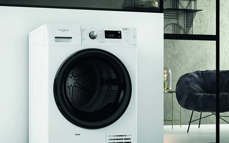 Un sèche-linge blanc Whirlpool avec un hublot noir est montré. Un fauteuil sombre est visible en arrière-plan.