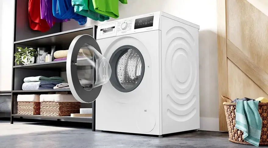 Witte wasmachine met open deur, rek met kleding en mand met wasgoed in een kamer.
