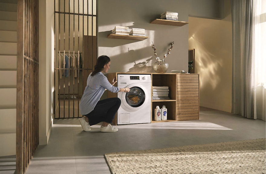 Une salle de lavage avec une machine à laver blanche, des étagères en bois, du linge et des produits de nettoyage.