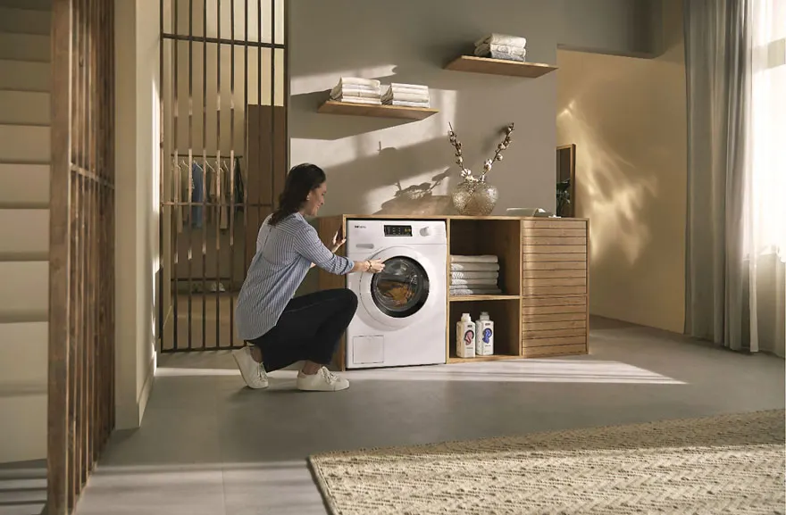 Une salle de lavage avec une machine à laver blanche, des étagères en bois, du linge et des produits de nettoyage.