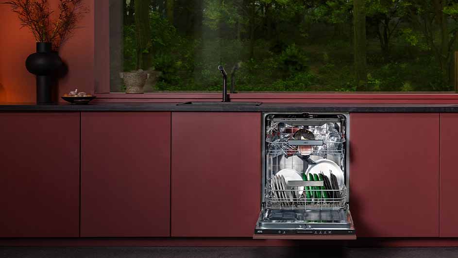 Open vaatwasser vol servies in een bordeauxrode keuken. Buiten een bos zichtbaar.