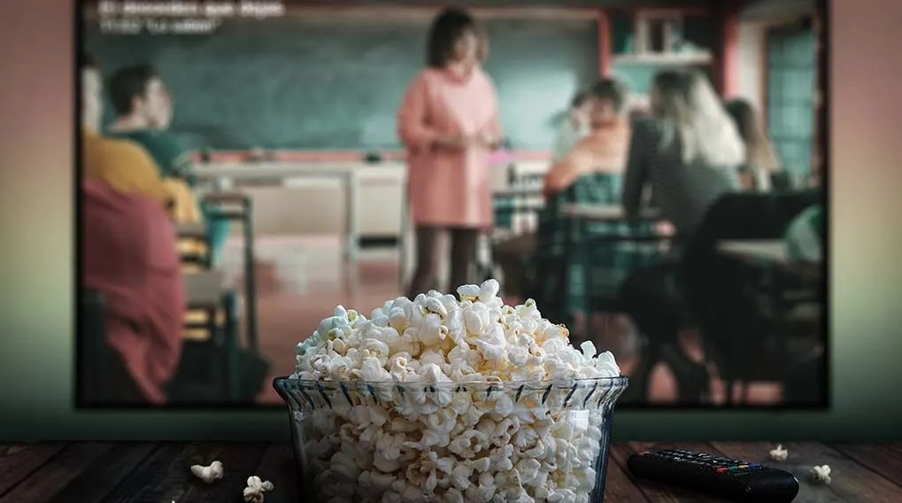 Bowl of popcorn in front of a TV showing a classroom scene, with a remote on a wood surface.