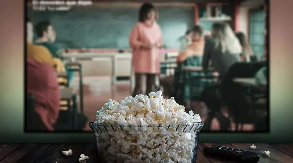 Bowl of popcorn in front of a TV showing a classroom scene, with a remote on a wood surface.