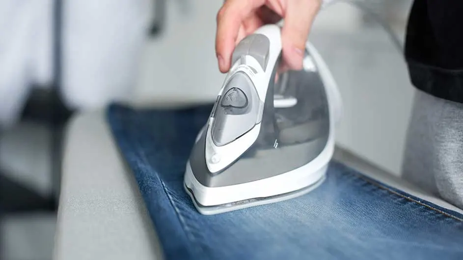 Een grijs en wit strijkijzer staat op een blauwe spijkerbroek. De achtergrond is licht en vaag.