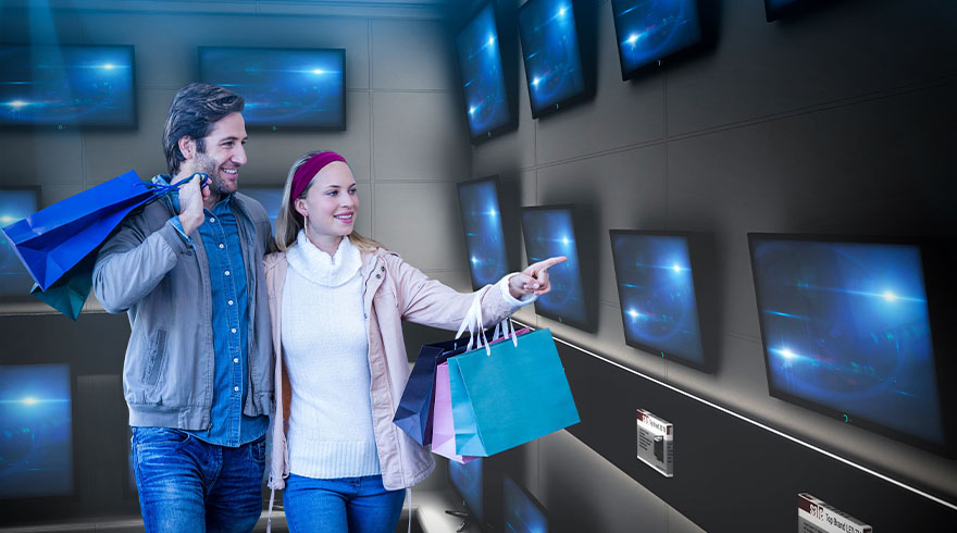 Een winkel met een wand vol televisies. Een vrouw wijst naar een scherm. Ze draagt een witte trui en een roze jas. Ze heeft boodschappentassen bij.