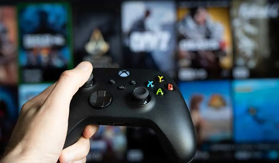 Une main tient une manette noire Xbox, avec des jeux Xbox flous visibles en arrière-plan sur un écran.