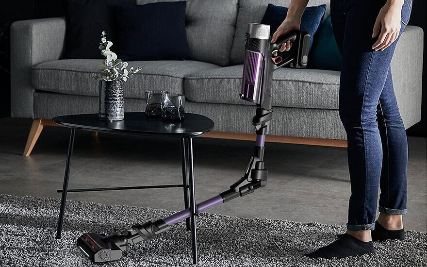 A cordless vacuum cleaner is on a grey rug in front of a black table and a grey couch.