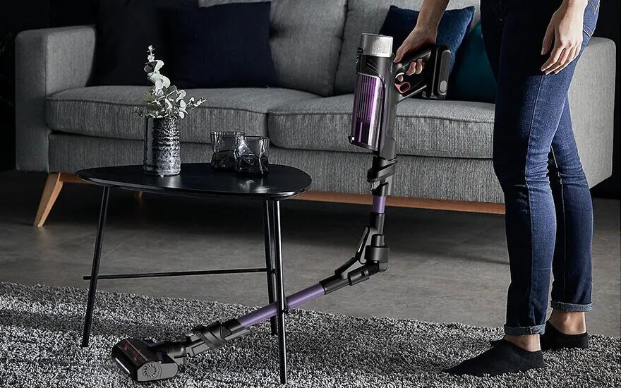 Un aspirateur noir et violet sur un tapis gris, près d'une table basse noire avec des décorations, devant un canapé gris.