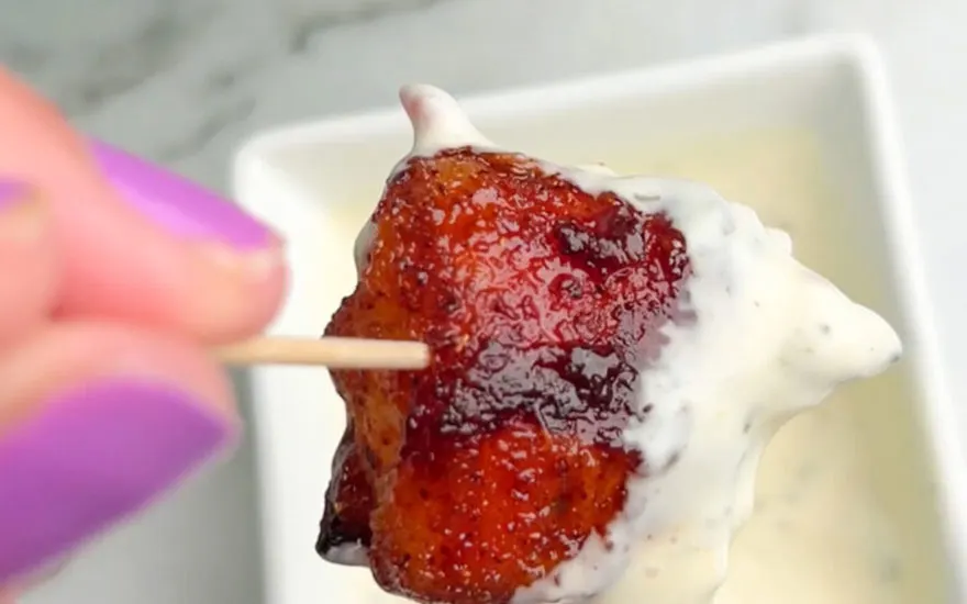 Gros plan d'un bonbon rouge sur un cure-dent, trempé dans une sauce blanche, devant un plat blanc.