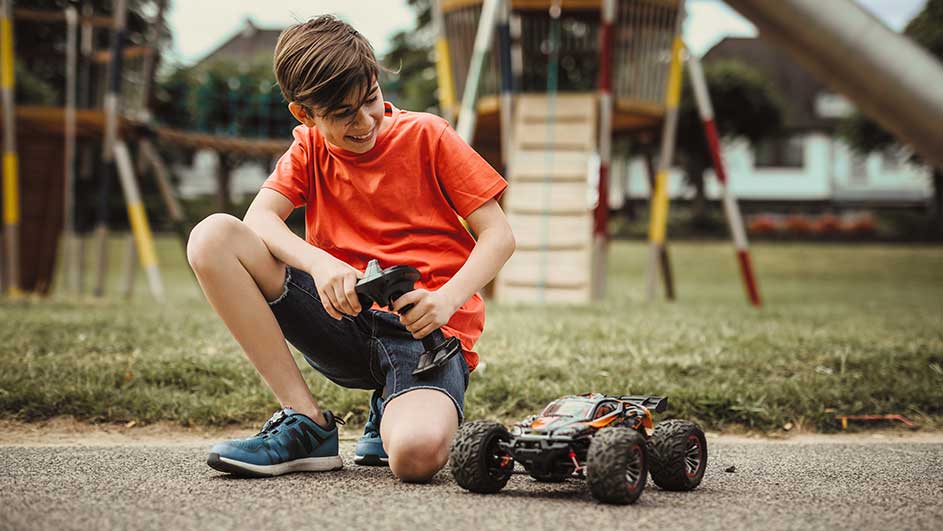 Een oranje speelgoedauto met afstandsbediening op een grijs pad, met een groene grasrand en een speeltuin op de achtergrond.