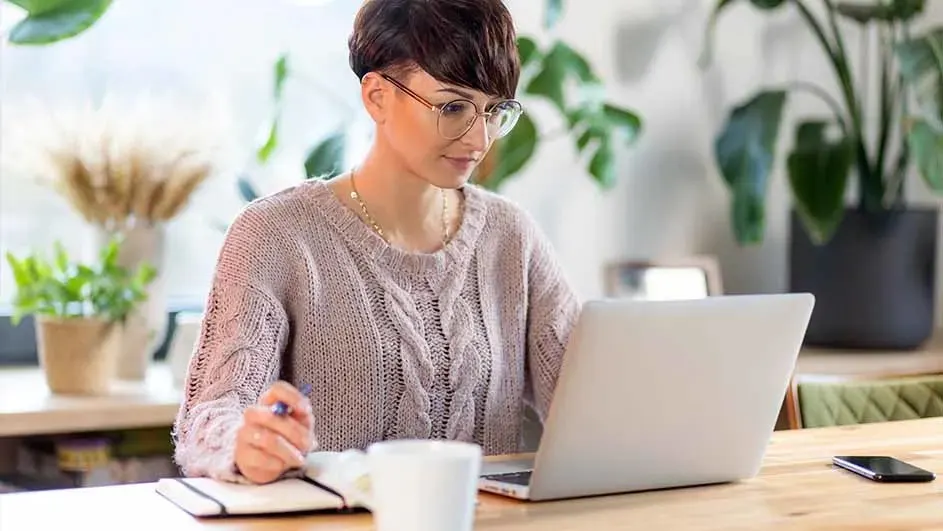 Une table en bois avec un ordinateur portable blanc, un bloc-notes, un stylo, une tasse blanche, un téléphone et des plantes en arrière-plan.