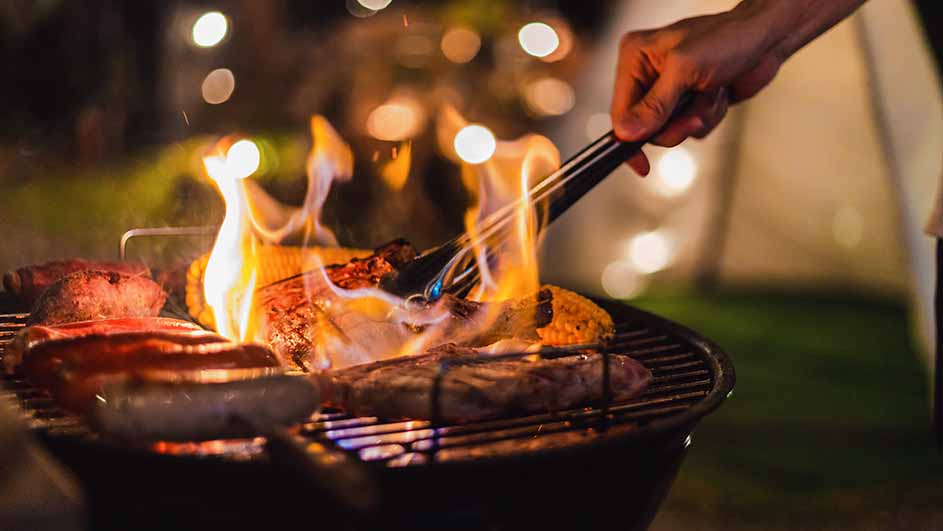 Un barbecue rond avec des flammes vives cuisant de la viande. Une main tient des pinces. Des lumières floues sont visibles à l'arrière-plan.