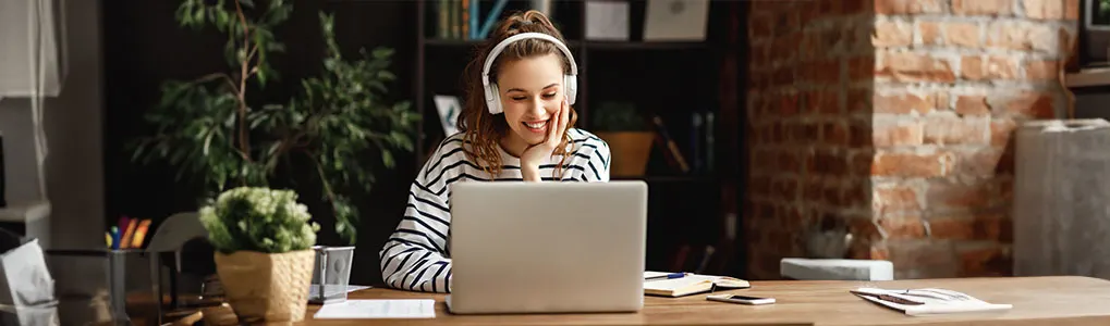Een laptop staat op een houten bureau met een notebook en een smartphone. Achteraan een bakstenen muur en een plant.