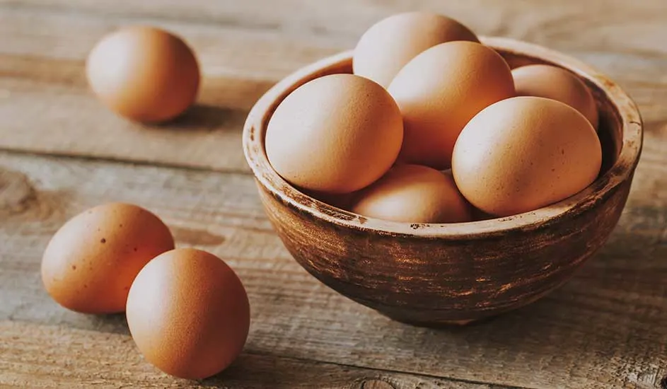 Un bol en bois rempli d'œufs bruns. Deux œufs supplémentaires sont placés à côté du bol sur une table en bois.