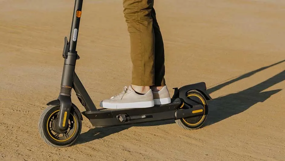 Een zwarte elektrische step staat op een zandkleurige ondergrond, met vage silhouetten. De step heeft gele accenten op de wielen.