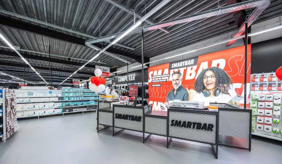 Interieur van een elektronicawinkel met een Smartbar-balie en rekken vol producten onder een metalen plafond met verlichting.