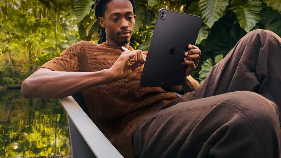 Un homme assis utilise une tablette noire avec un stylet dans un cadre extérieur avec des feuilles vertes.