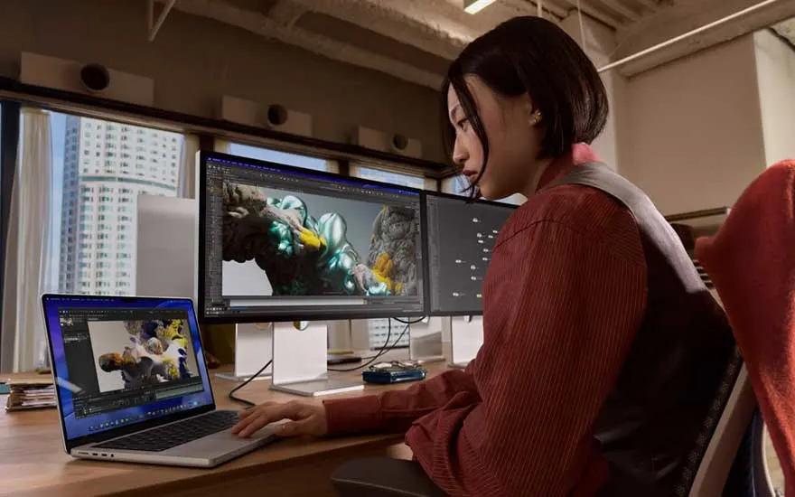 Een ontwerpster werkt met laptop en twee brede monitoren, waarop een kleurrijke digitale illustratie van een vogel wordt bewerkt.