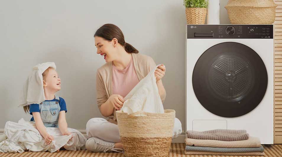 Une mère et son enfant rient près d’un panier à linge, à côté d’un sèche-linge moderne, avec serviettes pliées et placards en bois.