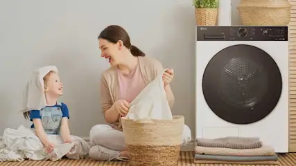Une mère et son enfant rient près d’un panier à linge, à côté d’un sèche-linge moderne, avec serviettes pliées et placards en bois.