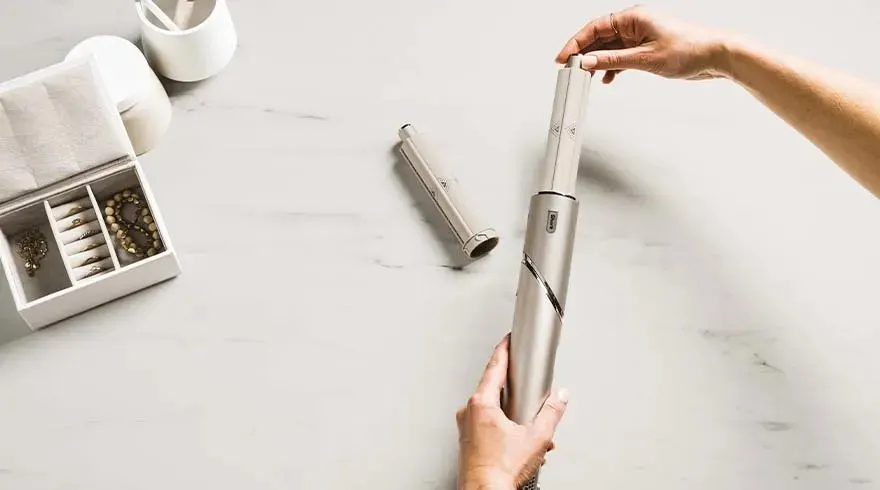 Sur une surface claire, une main assemble un fer à friser gris. Une boîte à bijoux ouverte et un pot blanc sont en arrière-plan.