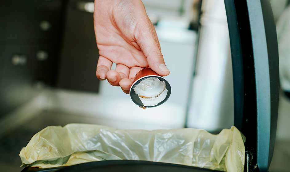 Een hand gooit een gebruikte koffiecapsule in een afvalbak met een gele vuilniszak, naast een open apparaat of kast in de keuken.