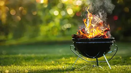 Een zwarte barbecue brandt met vlammen en rook op een gazon, met een onscherpe achtergrond van bomen en licht.