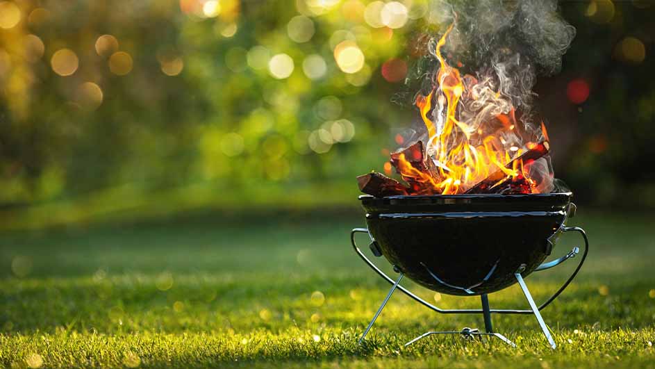 Un barbecue noir sur une pelouse verte. Des flammes et de la fumée s'élèvent du charbon de bois. L'arrière-plan est flou.