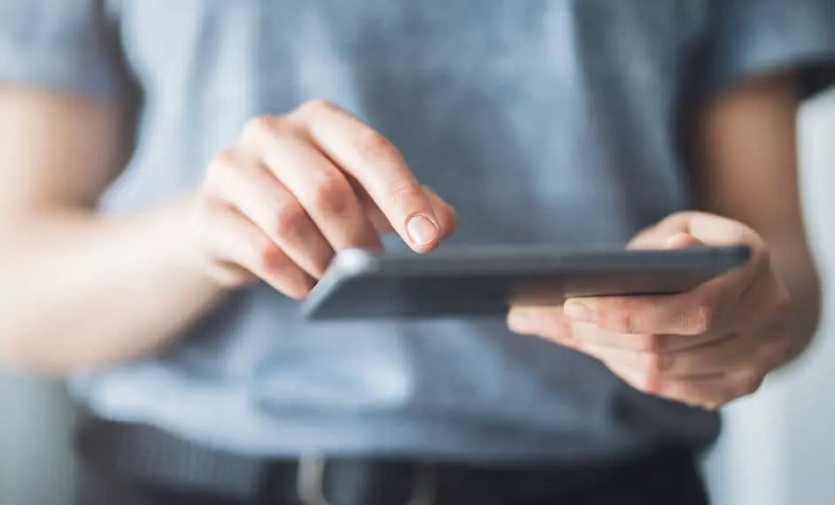 Gros plan des mains tenant une tablette noire. Un doigt appuie sur l'écran. Vêtements gris en arrière-plan.