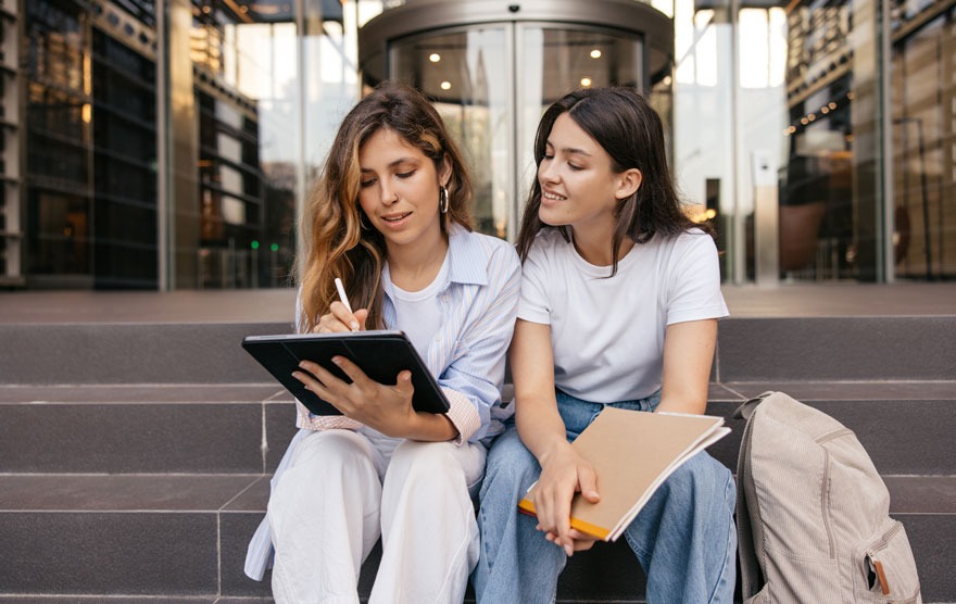 Twee personen zitten op een trap met een tablet en een notitieboek. Achter hen staat een gebouw met een draaideur.