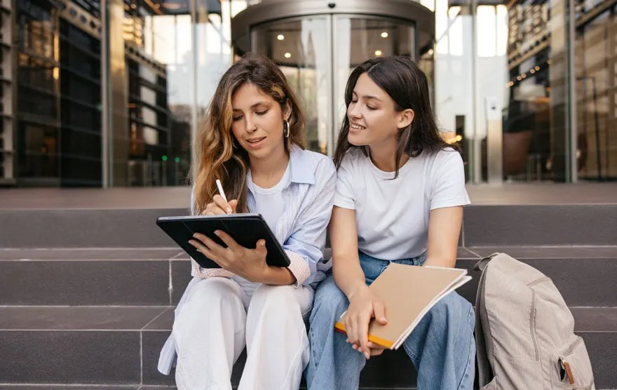 Twee personen zitten op een trap met een tablet en een notitieboek. Achter hen staat een gebouw met een draaideur.