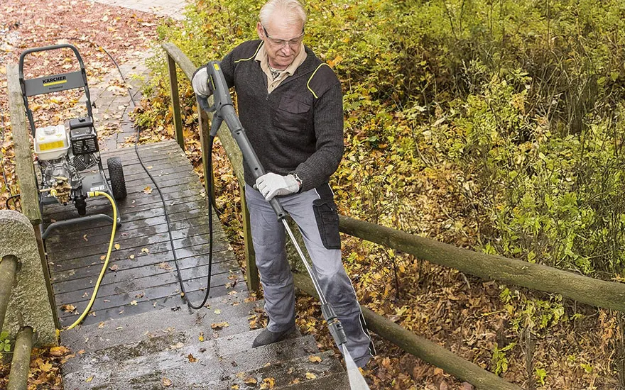 Een oudere man in donkere kleding reinigt een houten leuning op een brug met herfstbladeren, terwijl zijn hogedrukreiniger op de achtergrond staat.