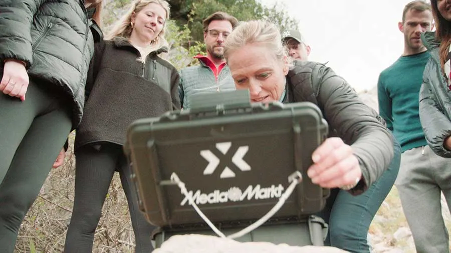 Een groep mensen kijkt naar een groene koffer van MediaMarkt, staand op een rotsachtige ondergrond. De achtergrond is licht bewolkt.