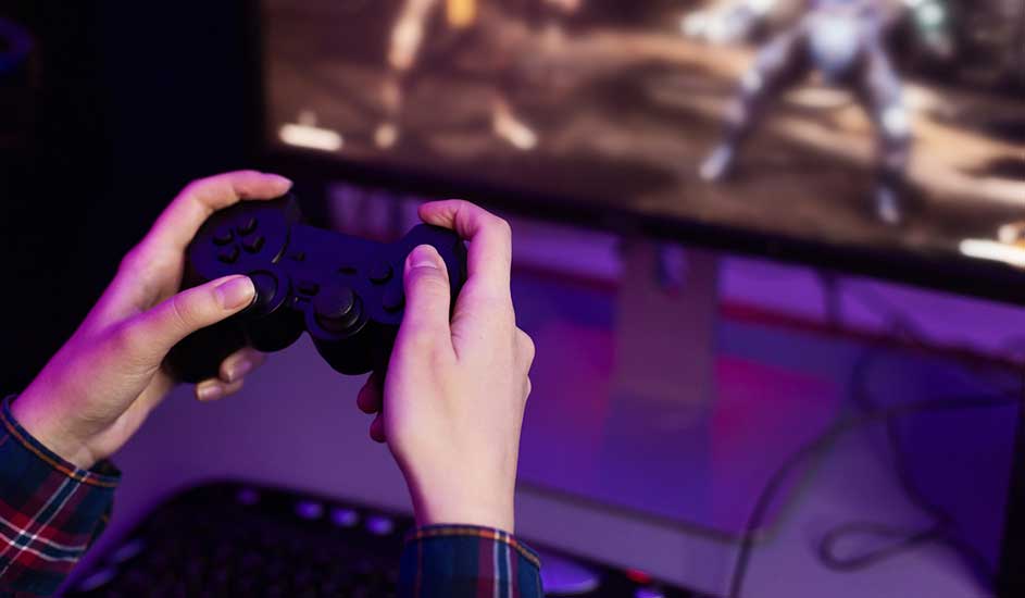 Une personne tient une manette noire devant un écran affichant un jeu vidéo aux tons violets et bruns. Un clavier est visible.