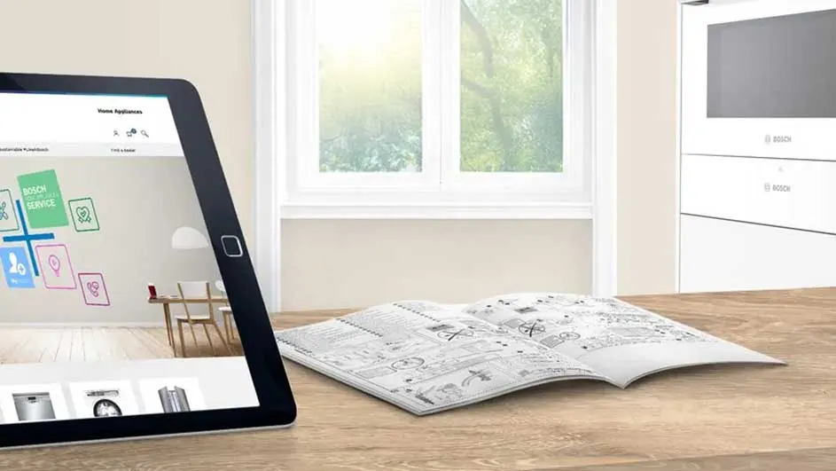 Een tablet, open handleiding en een witte kast staan op een houten tafel voor een raam.