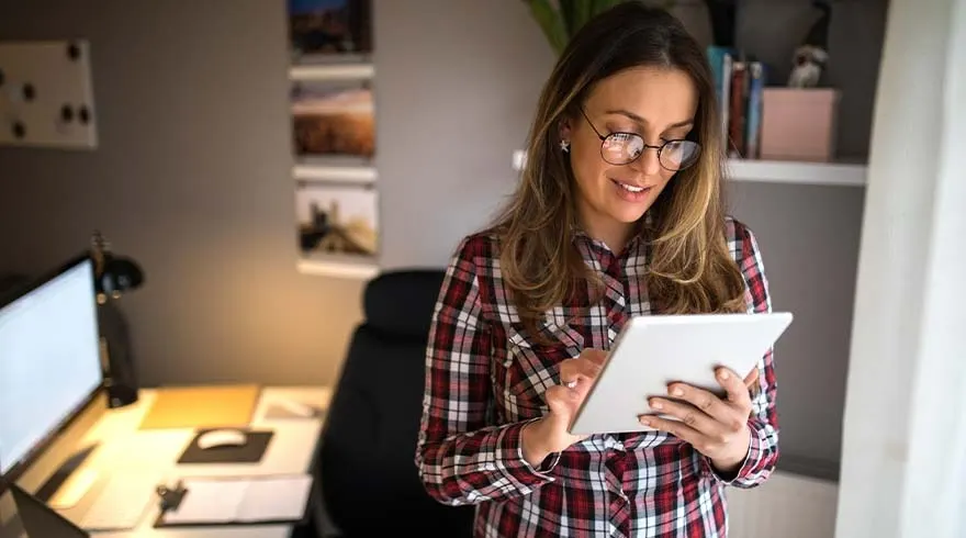 Une tablette blanche est tenue devant un bureau avec un ordinateur, des papiers et une lampe noire. Une étagère est visible en arrière-plan.