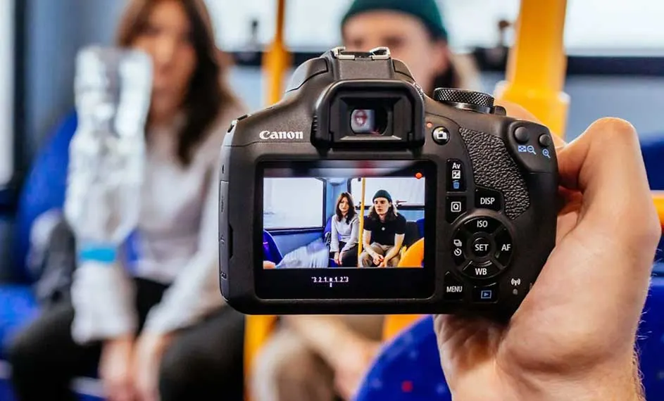 Un appareil photo Canon noir tient une photo de deux personnes assises dans un bus bleu et blanc.