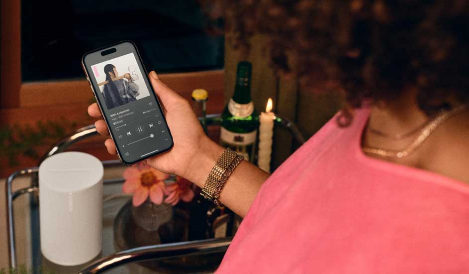 Hand houdt een smartphone met een muziek-app vast. Op een bar trolley staan een witte speaker, bloemen, een fles en een kaars.