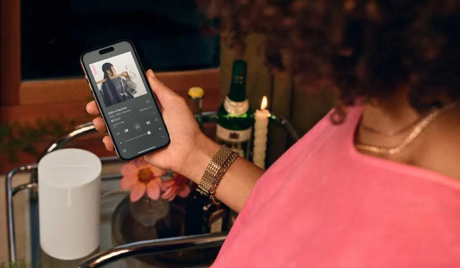Hand houdt een smartphone met een muziek-app vast. Op een bar trolley staan een witte speaker, bloemen, een fles en een kaars.