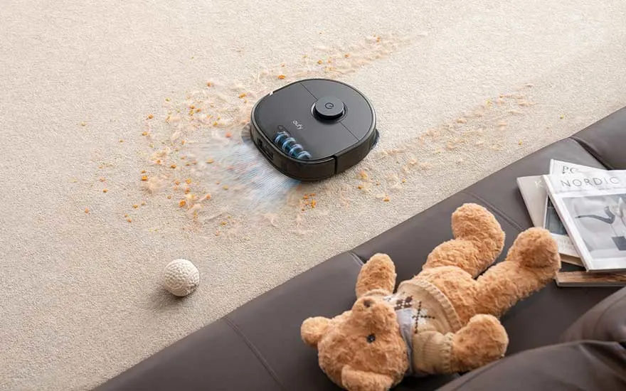 Een zwarte robotstofzuiger maakt een beige tapijt schoon bezaaid met kruimels. Naast de bank liggen een bal en een knuffelbeer.