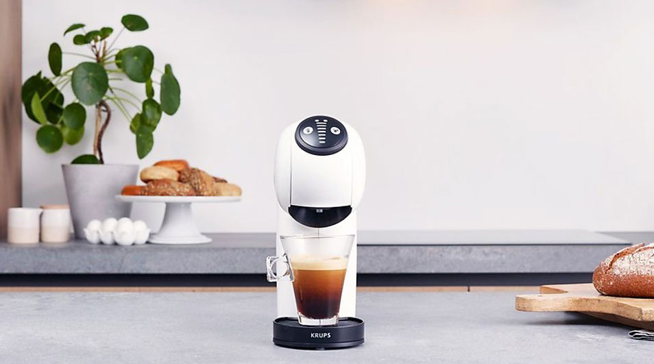 A white Krups coffee machine is brewing coffee into a glass. Behind it is a plant, pastry, and bread on a grey counter.