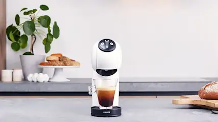 A white Krups coffee machine is brewing coffee into a glass. Behind it is a plant, pastry, and bread on a grey counter.