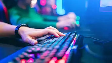 Une main tape sur un clavier rétroéclairé multicolore. Un poignet porte une montre. Un câble noir est visible.