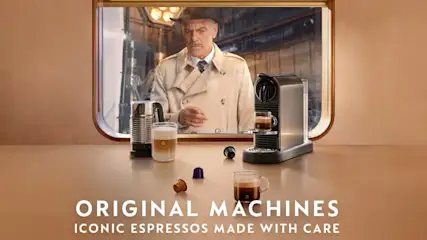 Man in beige regenjas kijkt verlangend door treinraam naar dampende espresso en capsules naast een compacte koffiemachine.