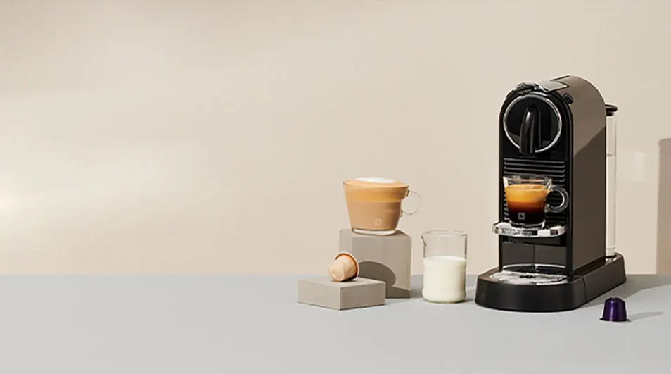 Machine à café noire avec tasse, pot à lait et capsule sur fond beige clair.